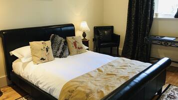 A neatly made bed with a white comforter and several decorative pillows, a black leather headboard, a small bedside table with a lamp, a black chair, and a window with patterned curtains.