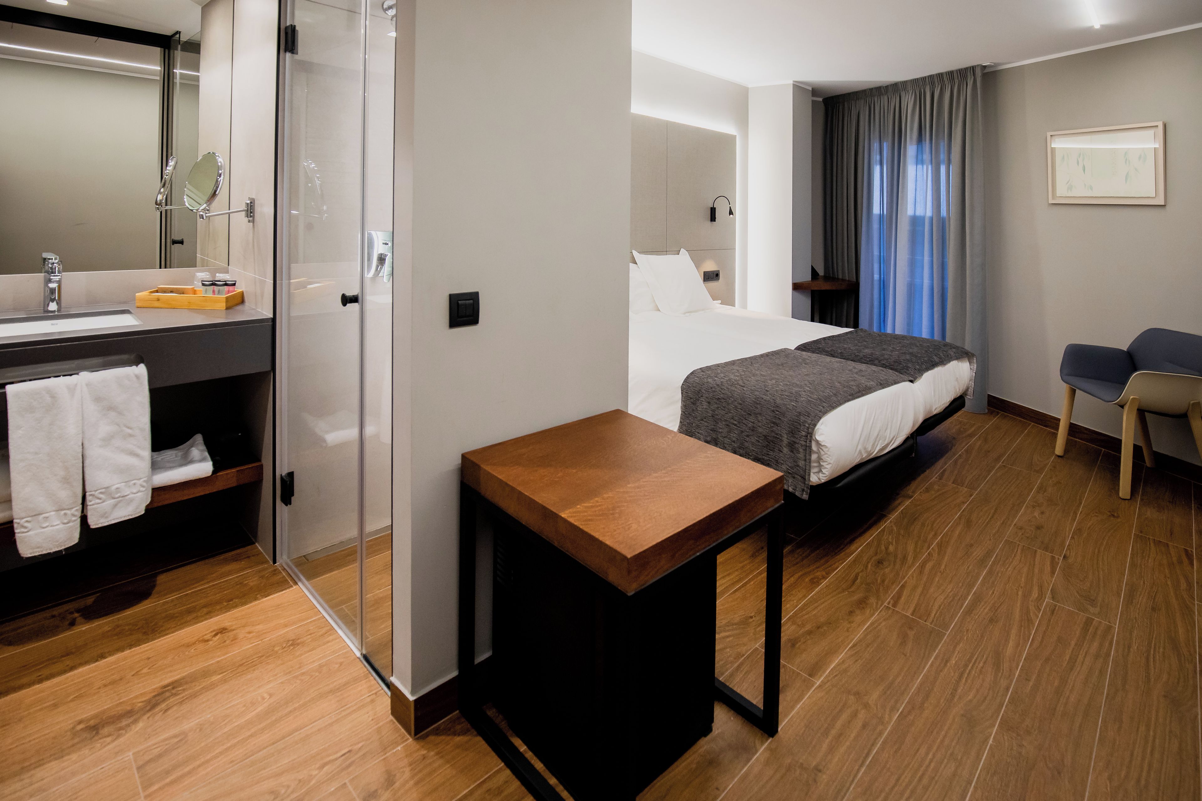 Habitación de hotel con piso de madera, una cama con ropa blanca, una mesita de noche, una pequeña mesa de madera, un baño con espejo, un perchero para toallas y una zona de ducha.