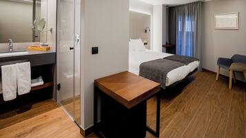 Habitación de hotel con piso de madera, una cama con ropa blanca, una mesita de noche, una pequeña mesa de madera, un baño con espejo, un perchero para toallas y una zona de ducha.