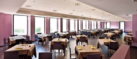Un comedor espacioso con varias mesas y sillas, ventanas amplias y un techo alto con iluminación empotrada.
