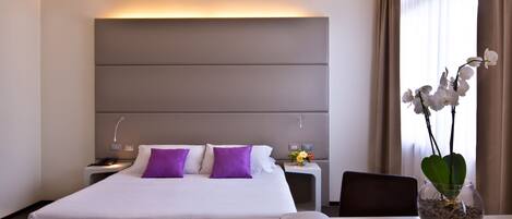 Una habitación de hotel con una cama grande, una mesita de noche con una lámpara, una pequeña mesa con un libro y un jarrón con flores.