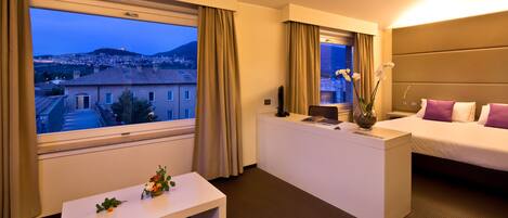 Una habitación de hotel con cama, escritorio, una pequeña mesa con flores, un jarrón y vistas a un paisaje urbano a través de ventanas amplias.