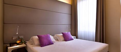 Una habitación de hotel moderna con una cama grande, dos mesitas de noche y un velador con lámpara, además de un ventanal con cortinas.
