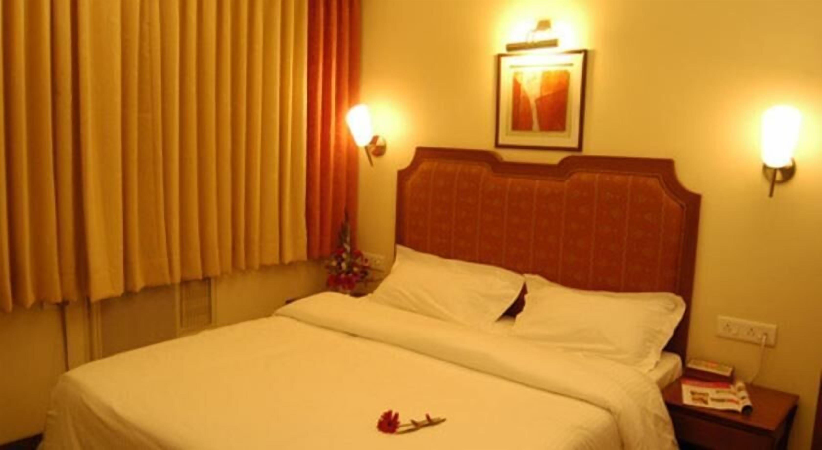 A hotel room with a bed, a nightstand, a wall-mounted lamp, and a window with curtains.
