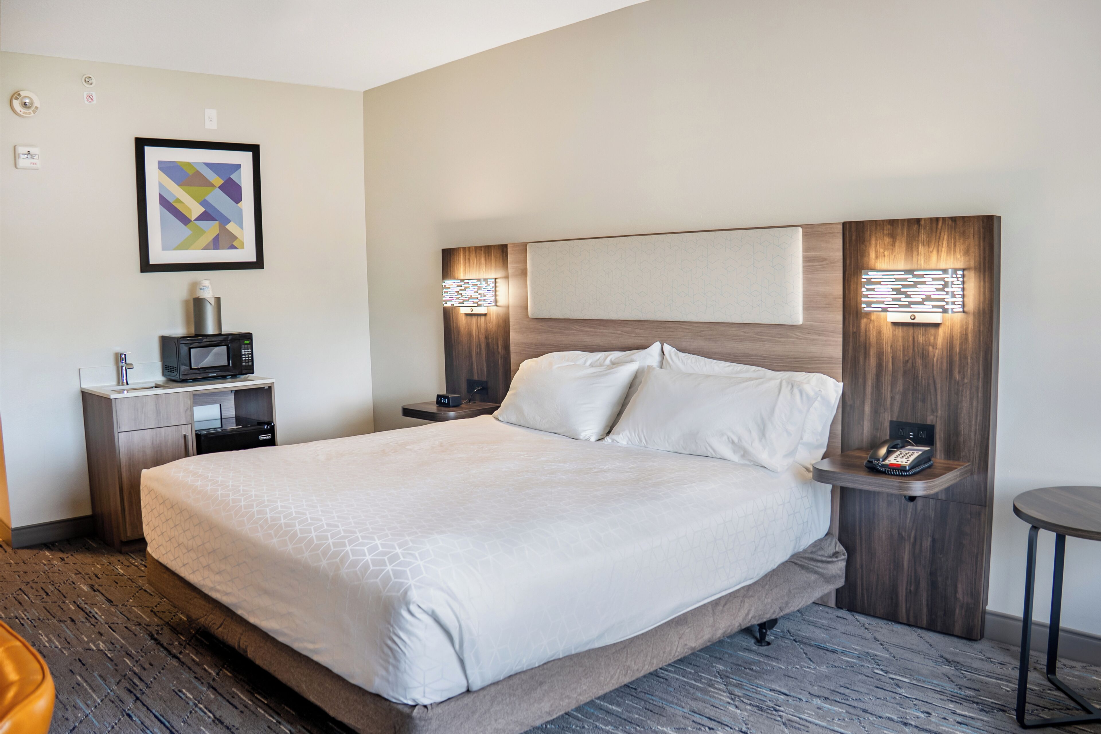 A hotel room with a large bed, a bedside table with a phone, a small desk with a microwave, and a framed picture on the wall.