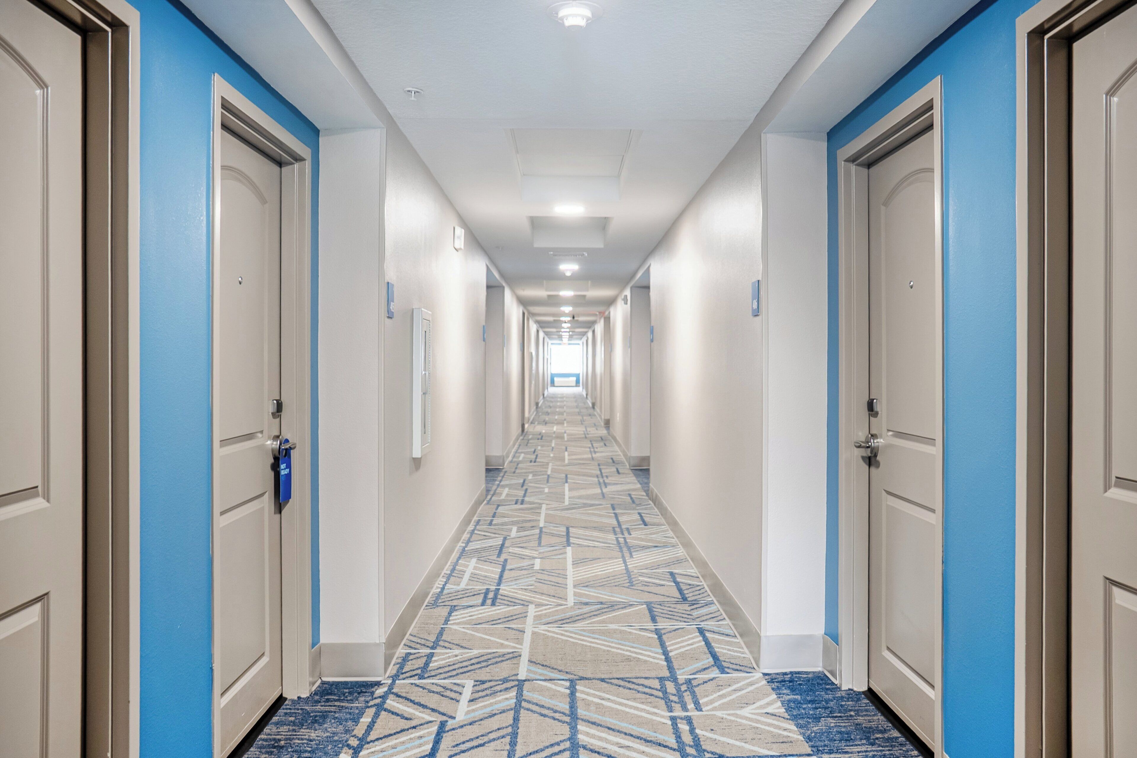 A hotel hallway with a patterned carpet, doors on both sides, and a light-colored ceiling.