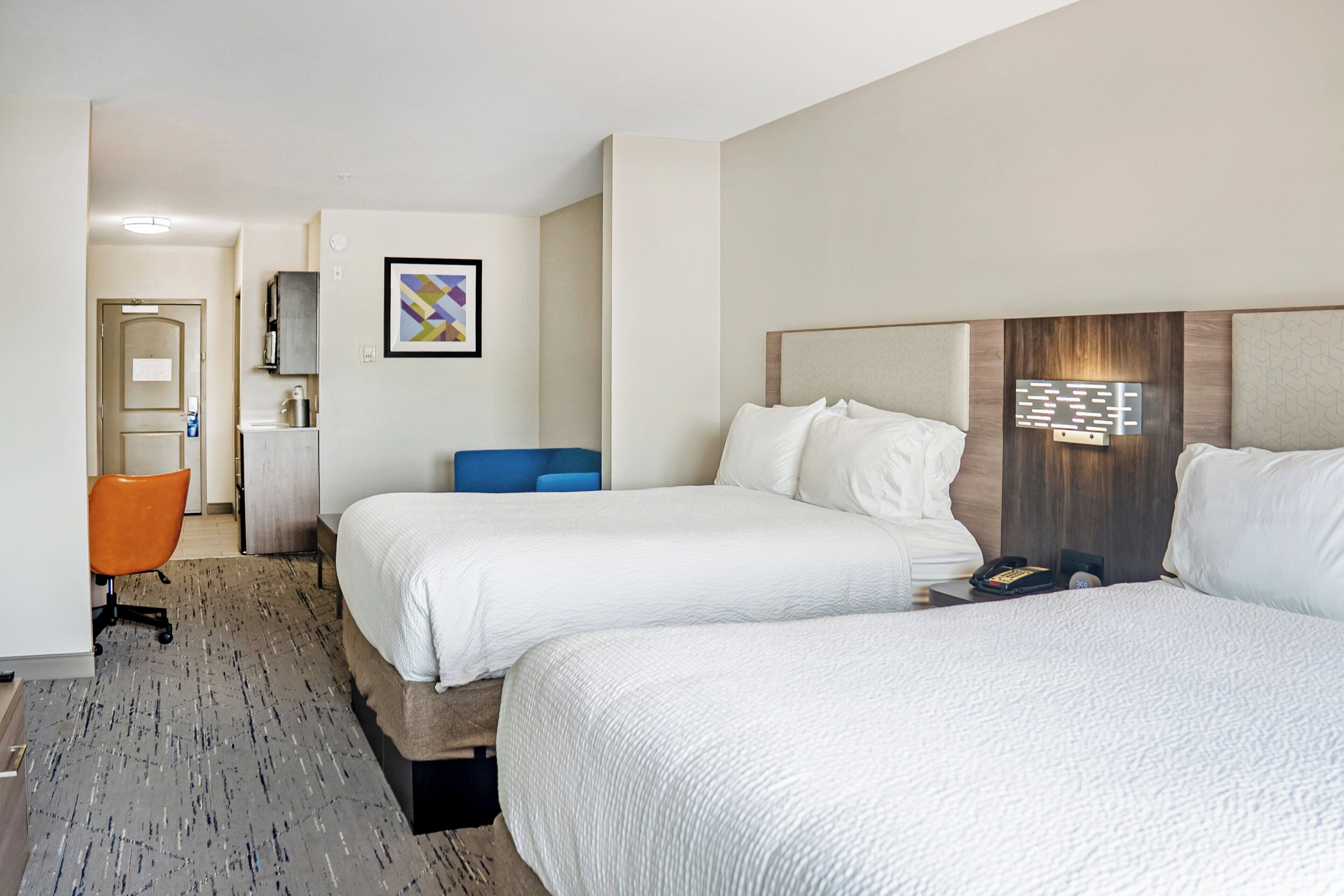 A hotel room with two beds, a desk, a chair, a small kitchenette, and a bathroom.