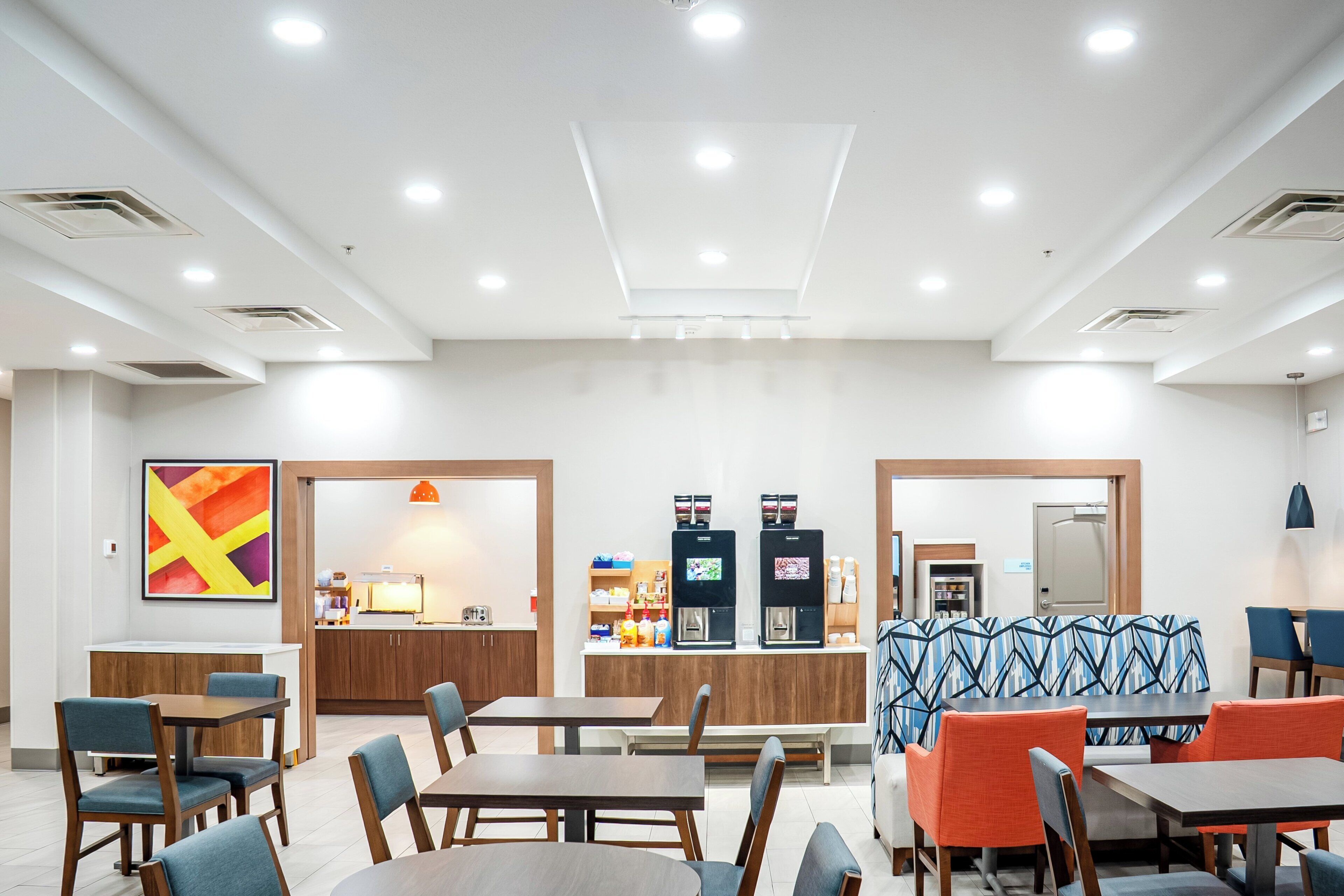 Un espace de restauration dans l’hôtel, avec des tables et des chaises, un coin café et un grand tableau abstrait accroché au mur.