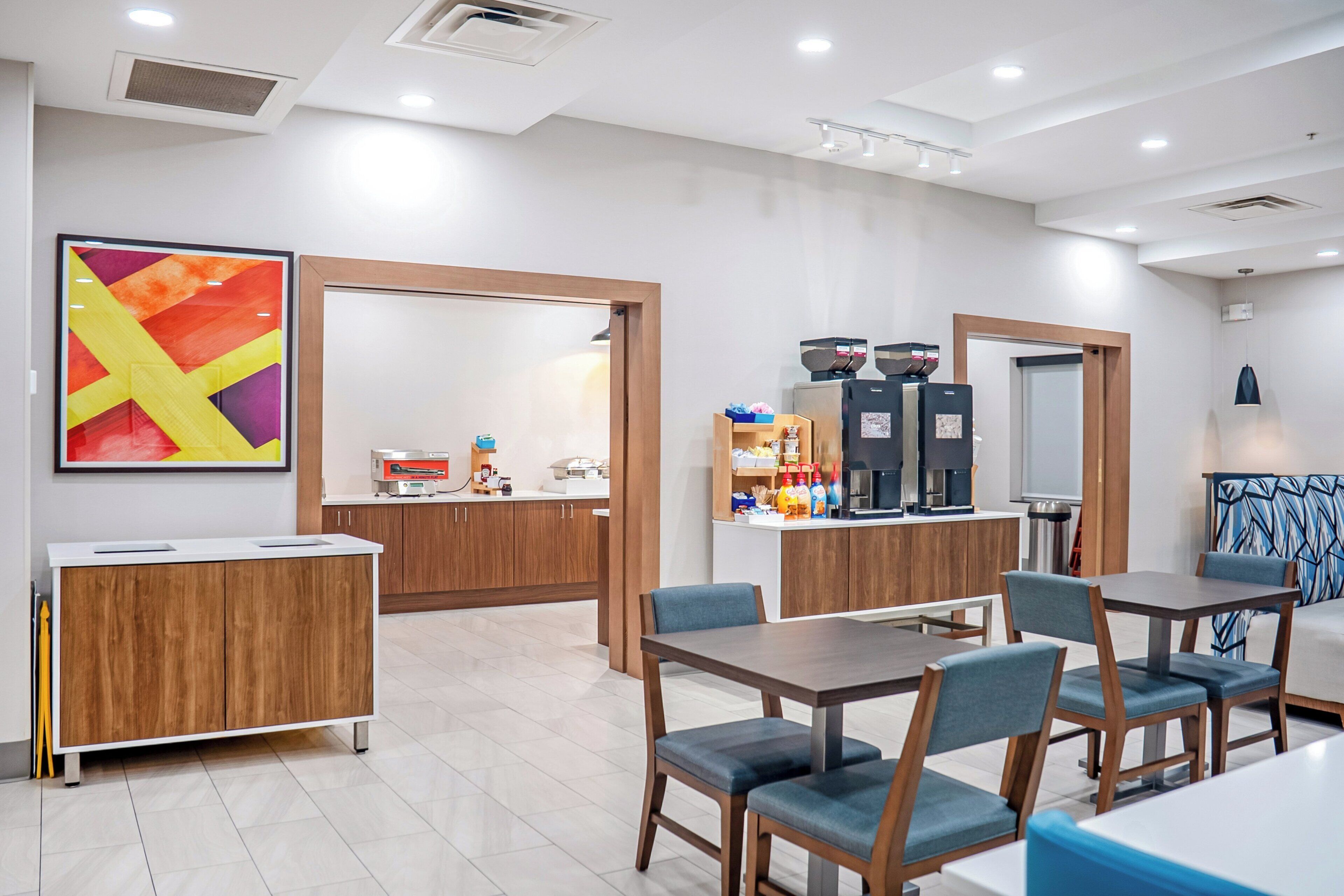 A modern hotel room with a kitchenette, a dining area with tables and chairs, and a large abstract painting on the wall.