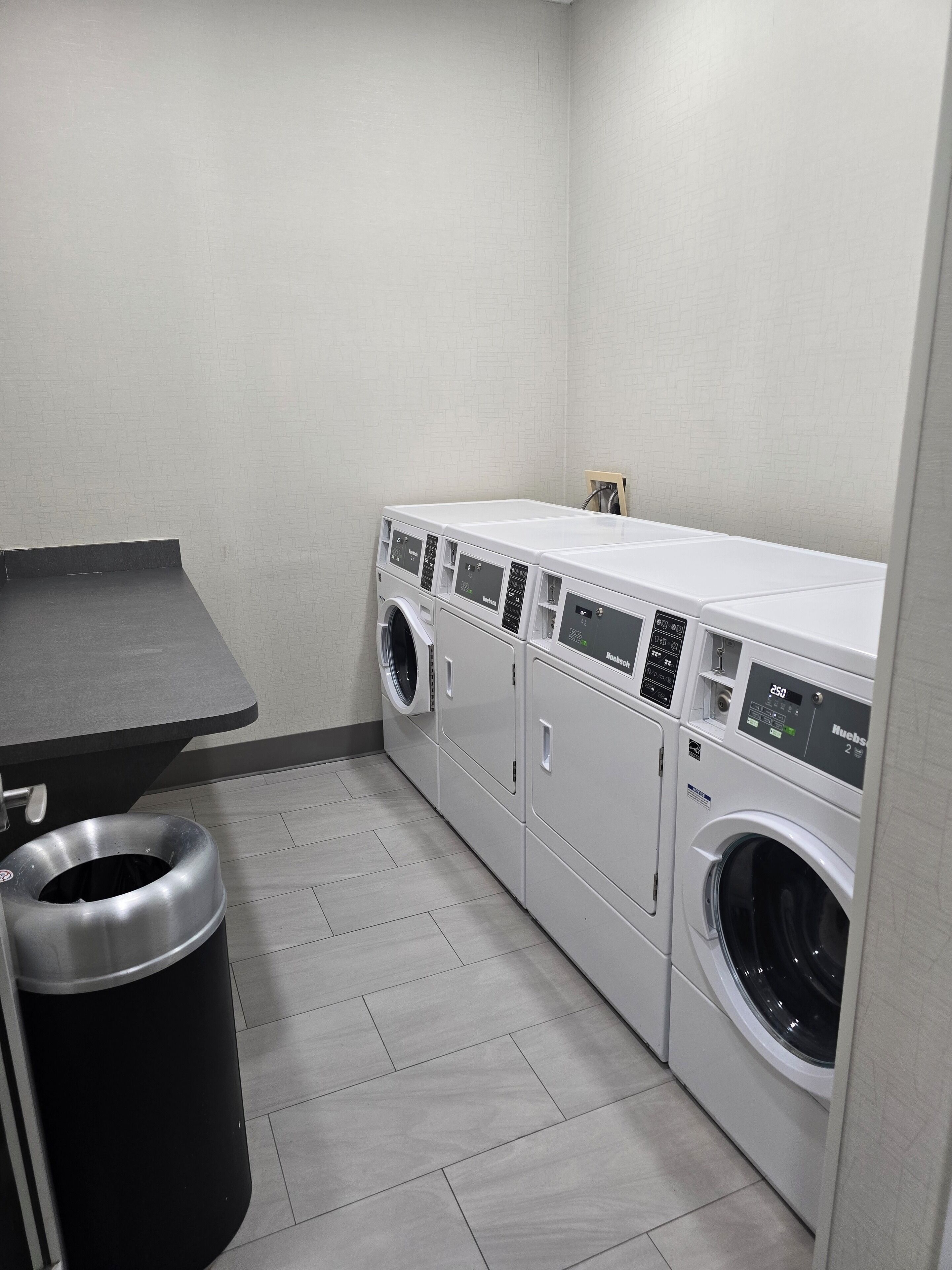 Une buanderie avec une rangée de lave-linge et de sèche-linge à chargement frontal blancs, un plan de travail gris et une poubelle noire.