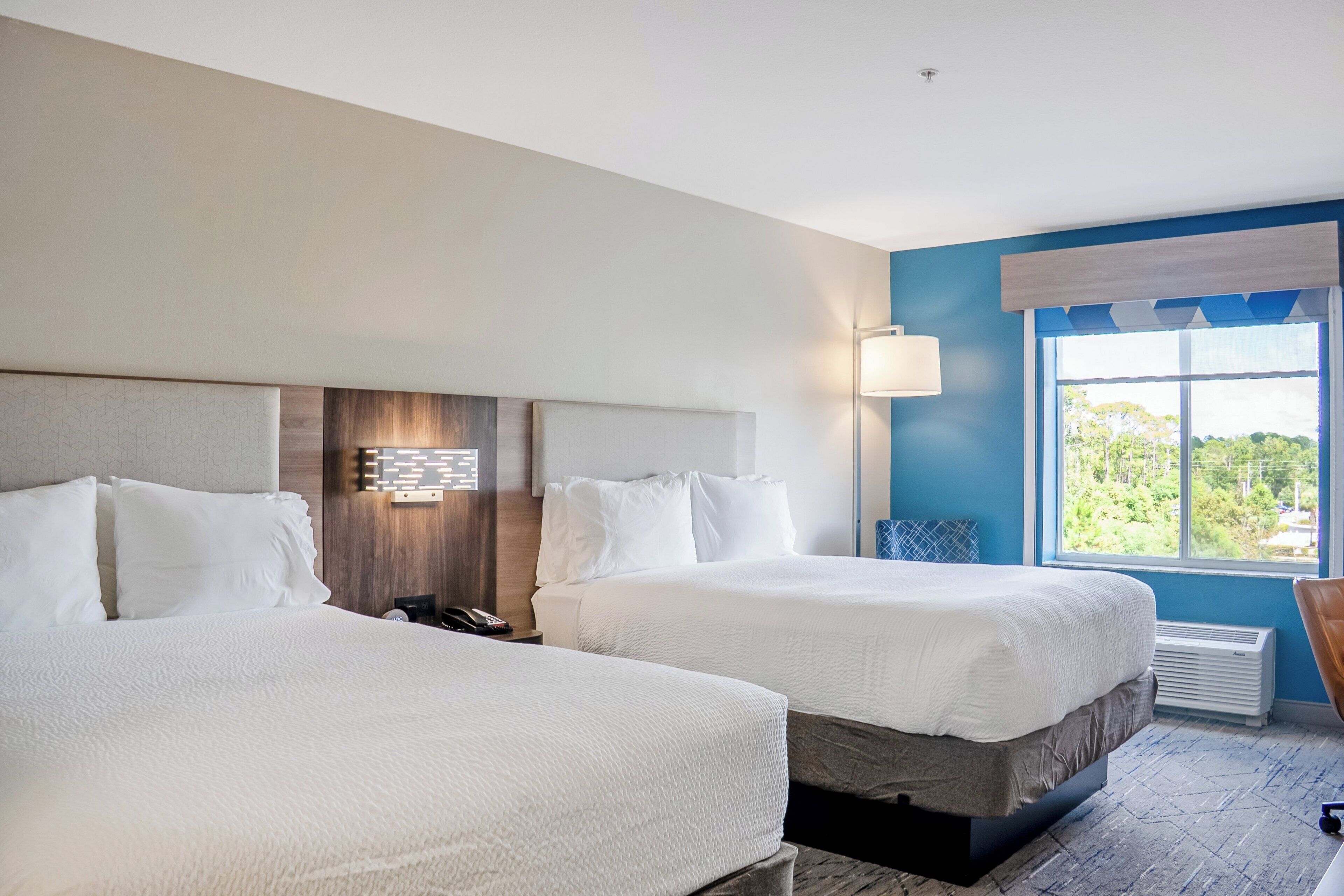 Een hotelkamer met twee bedden, een nachtkastje met een lamp, en een raam met uitzicht op bomen.