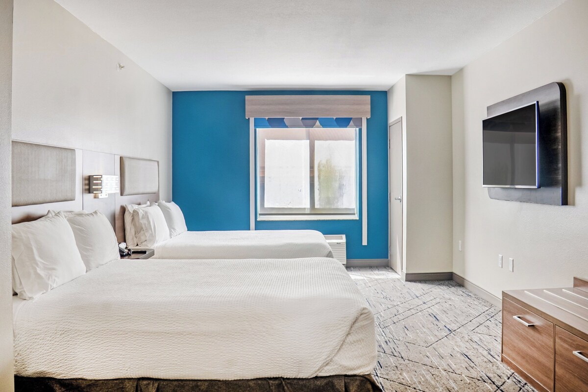 A hotel room with two beds, a large window with a view, a flat-screen TV mounted on the wall, and a wooden dresser.
