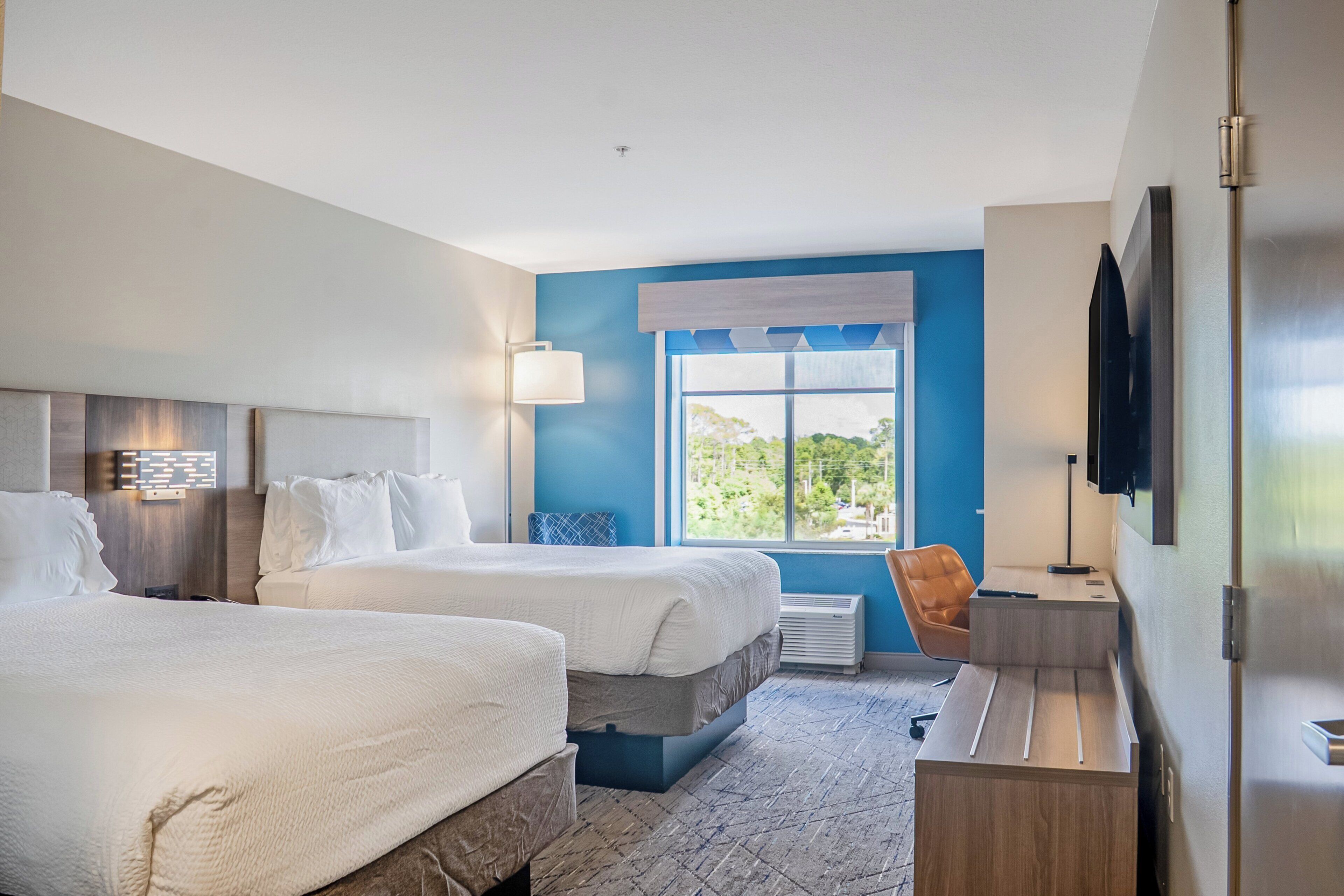 A hotel room with two beds, a desk, a chair, a TV, and a window with a view of greenery.