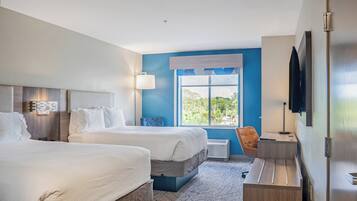 A hotel room with two beds, a desk, a chair, a TV, and a window with a view of greenery.