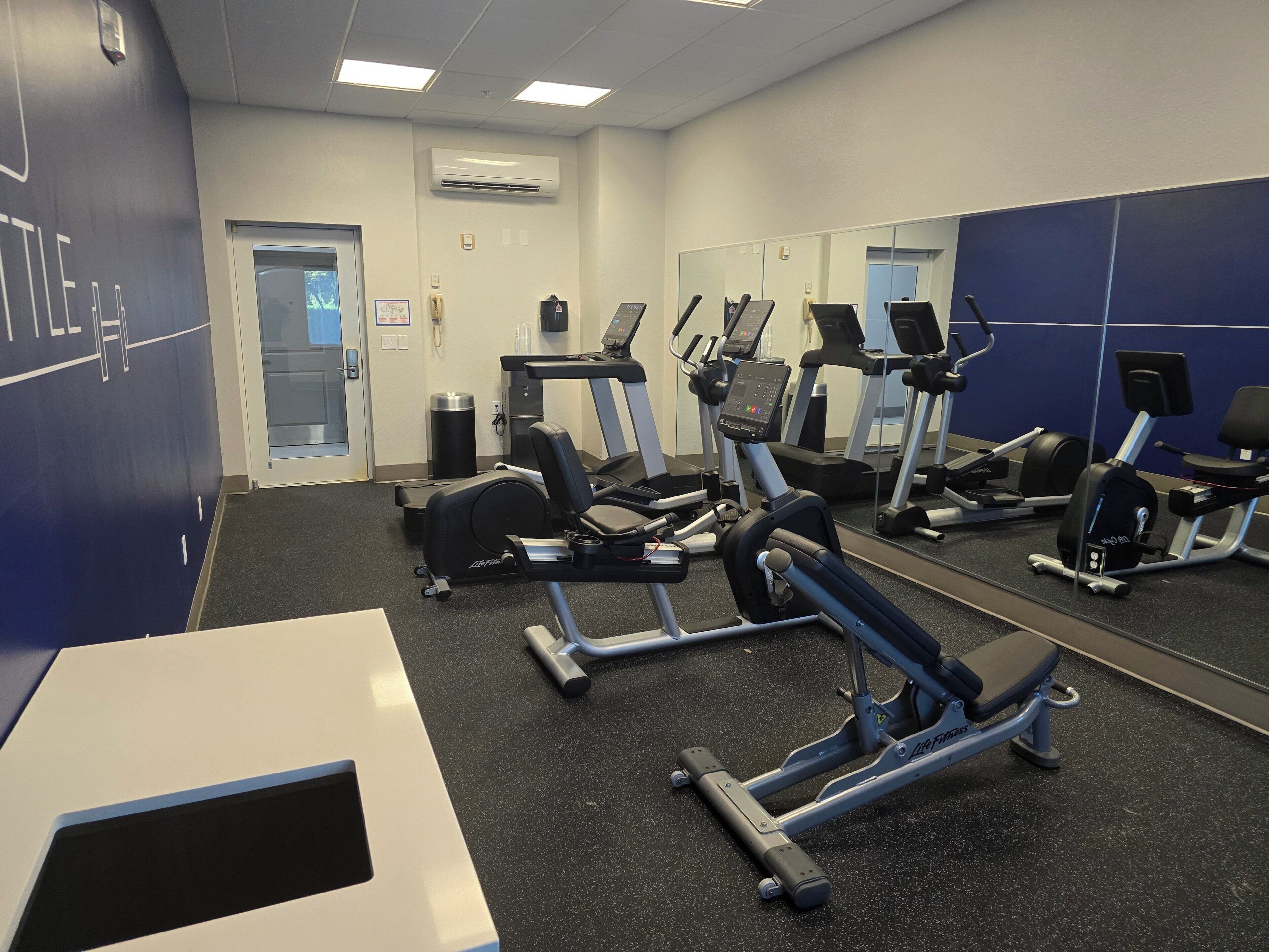 Un gimnasio con máquinas de cardio, un muro de espejos y una puerta que da a otra sala.