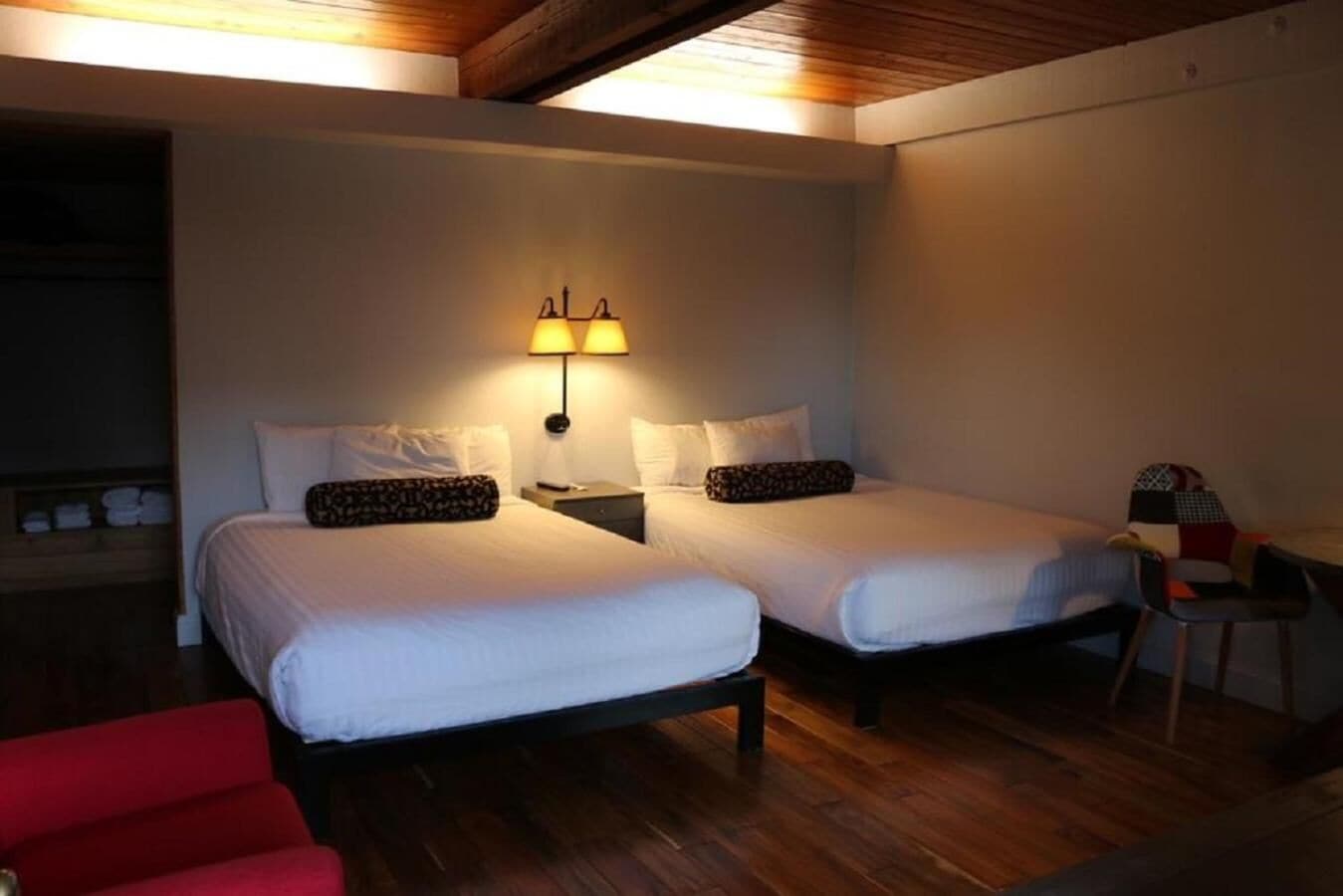 A room with two beds, a nightstand with a lamp, a red chair, and a wooden ceiling.
