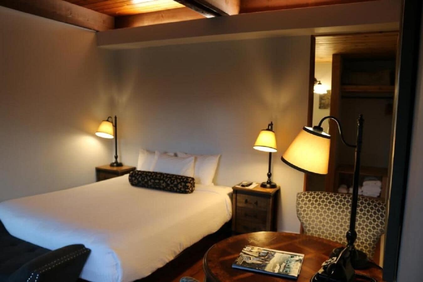 A hotel room with a bed, two bedside tables with lamps, a small round table with a magazine, and a patterned chair.