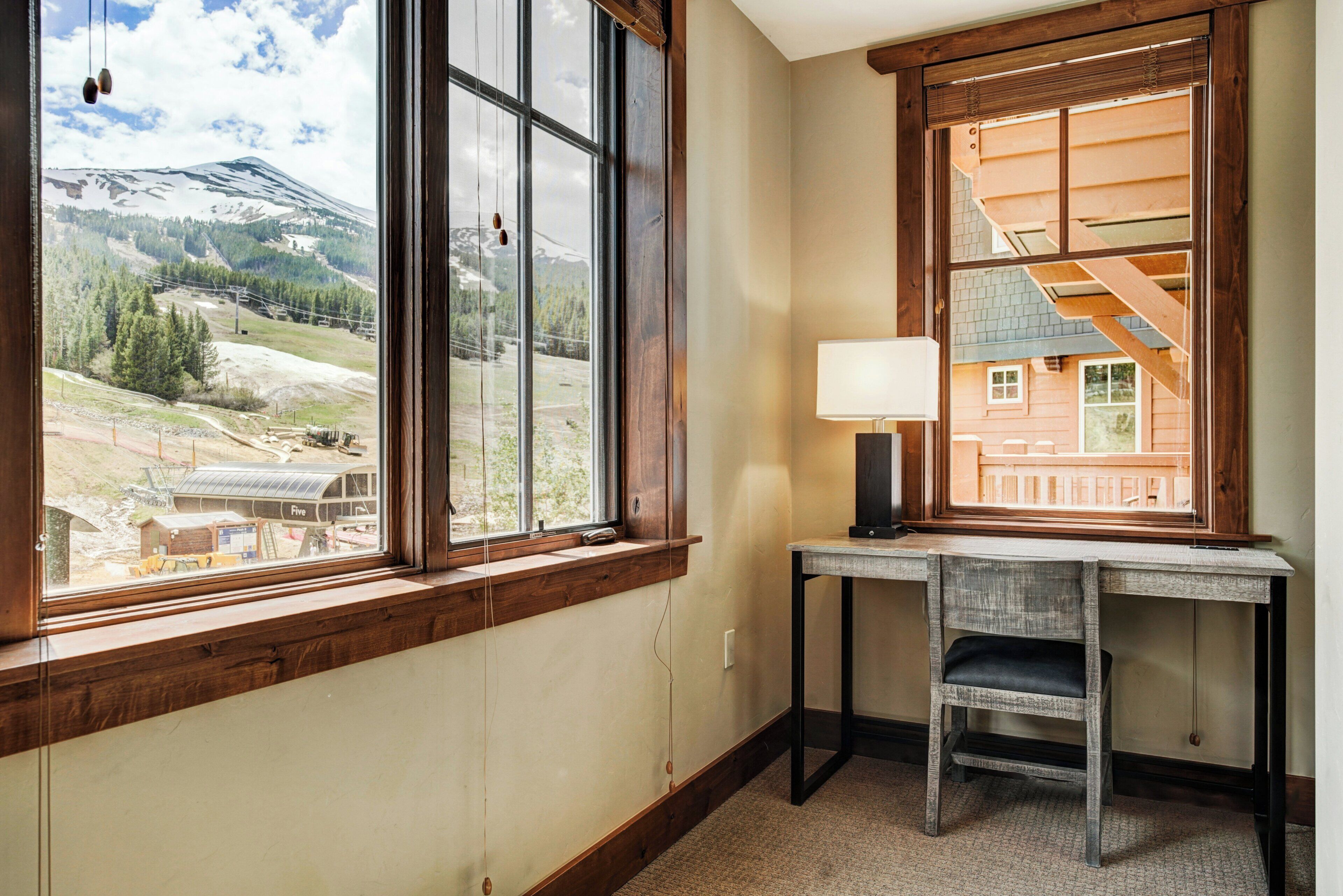 A room with a large window offering a view of a mountainous landscape, a desk with a chair, and a lamp.