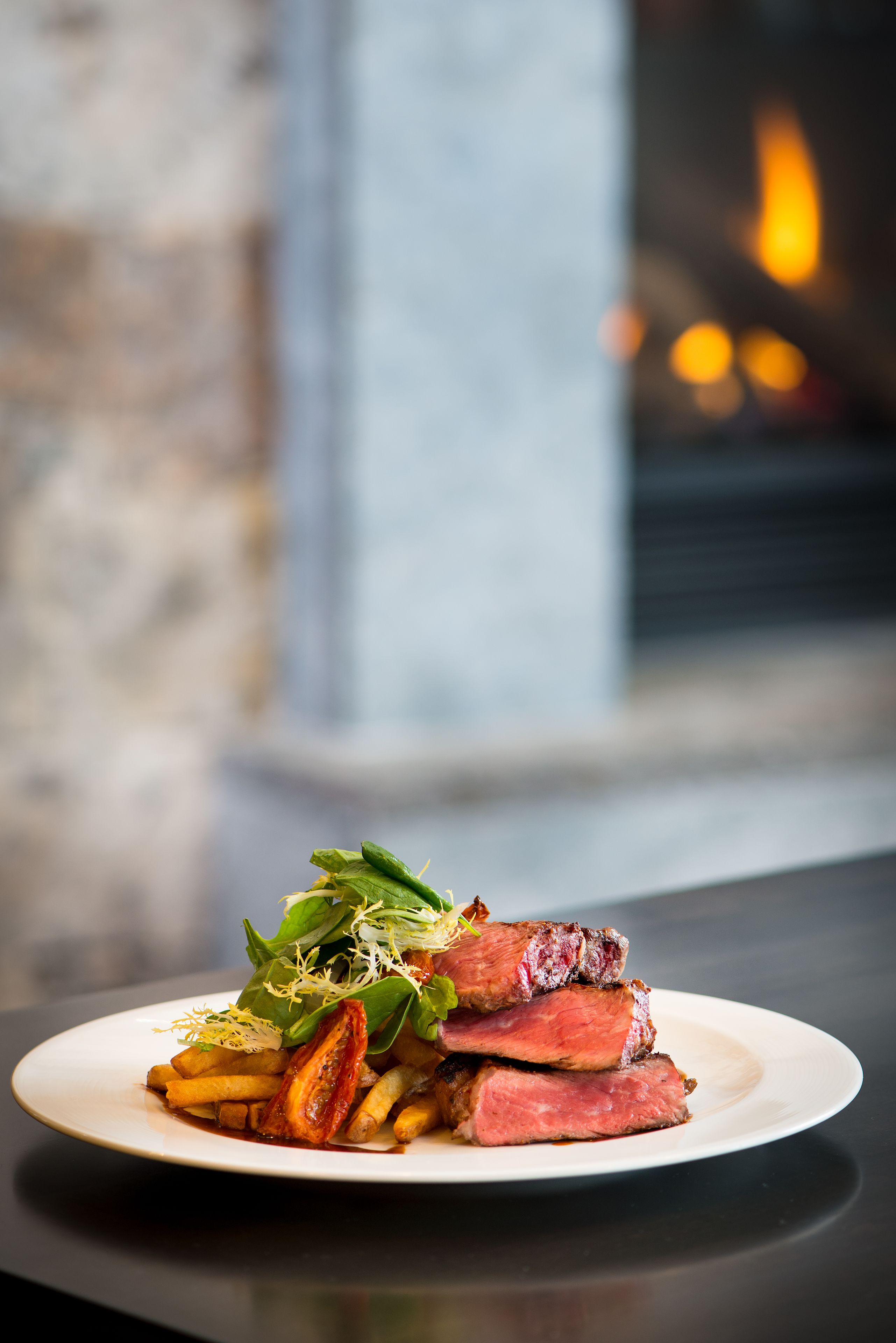 A plate of steak with fries and greens on a table with a blurred background of a fireplace.
