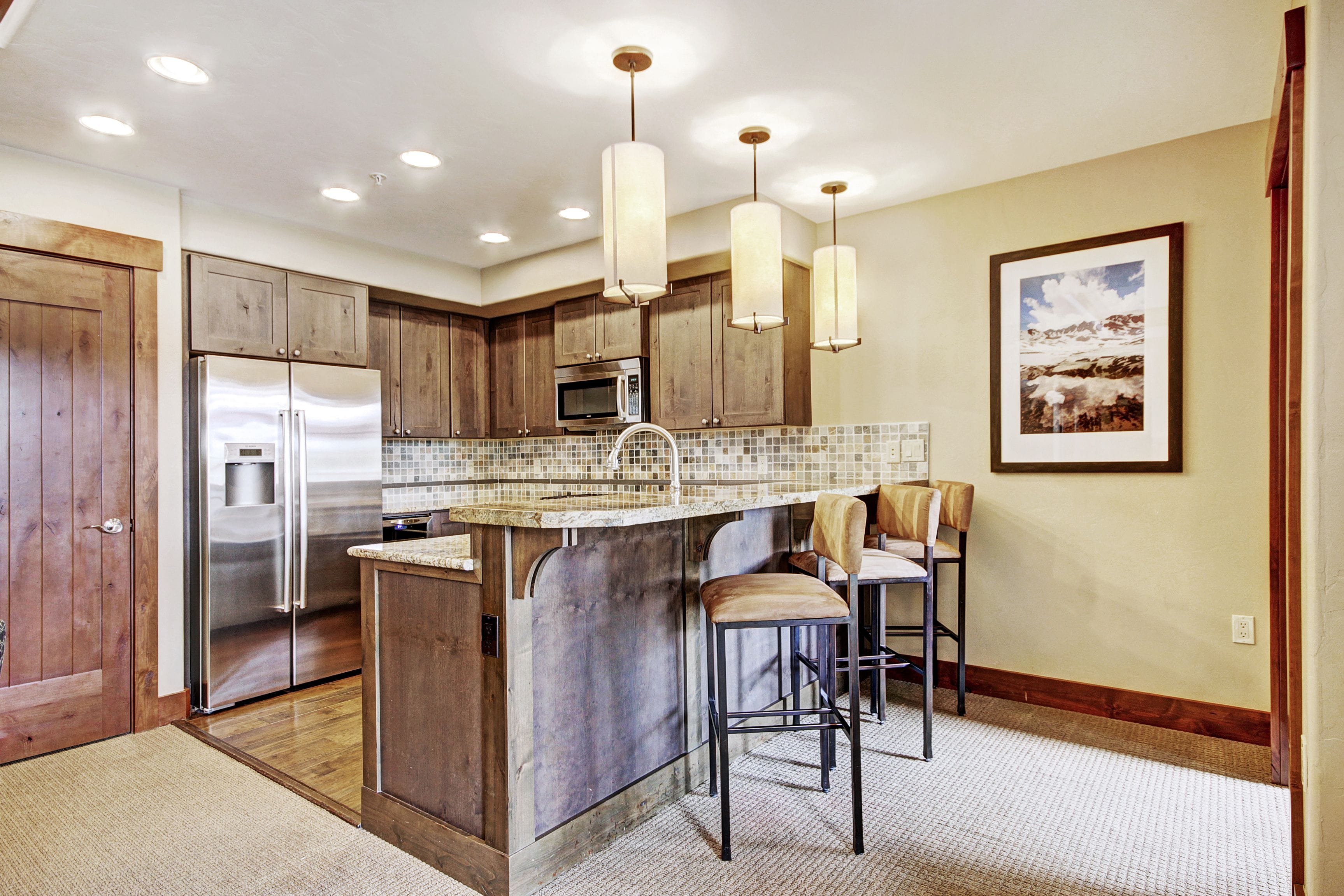 A modern kitchen with wooden cabinets, stainless steel appliances, a central island with bar stools, pendant lighting, and a framed landscape picture on the wall.