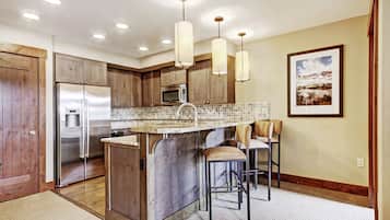 A modern kitchen with wooden cabinets, stainless steel appliances, a central island with bar stools, pendant lighting, and a framed landscape picture on the wall.