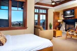 A room with a bed, a sofa, a TV mounted above a fireplace, and a view of mountains through large windows.