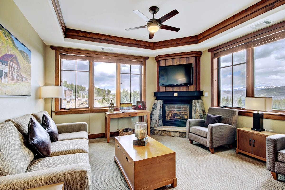 A cozy living room with a fireplace, large windows, and a ceiling fan.