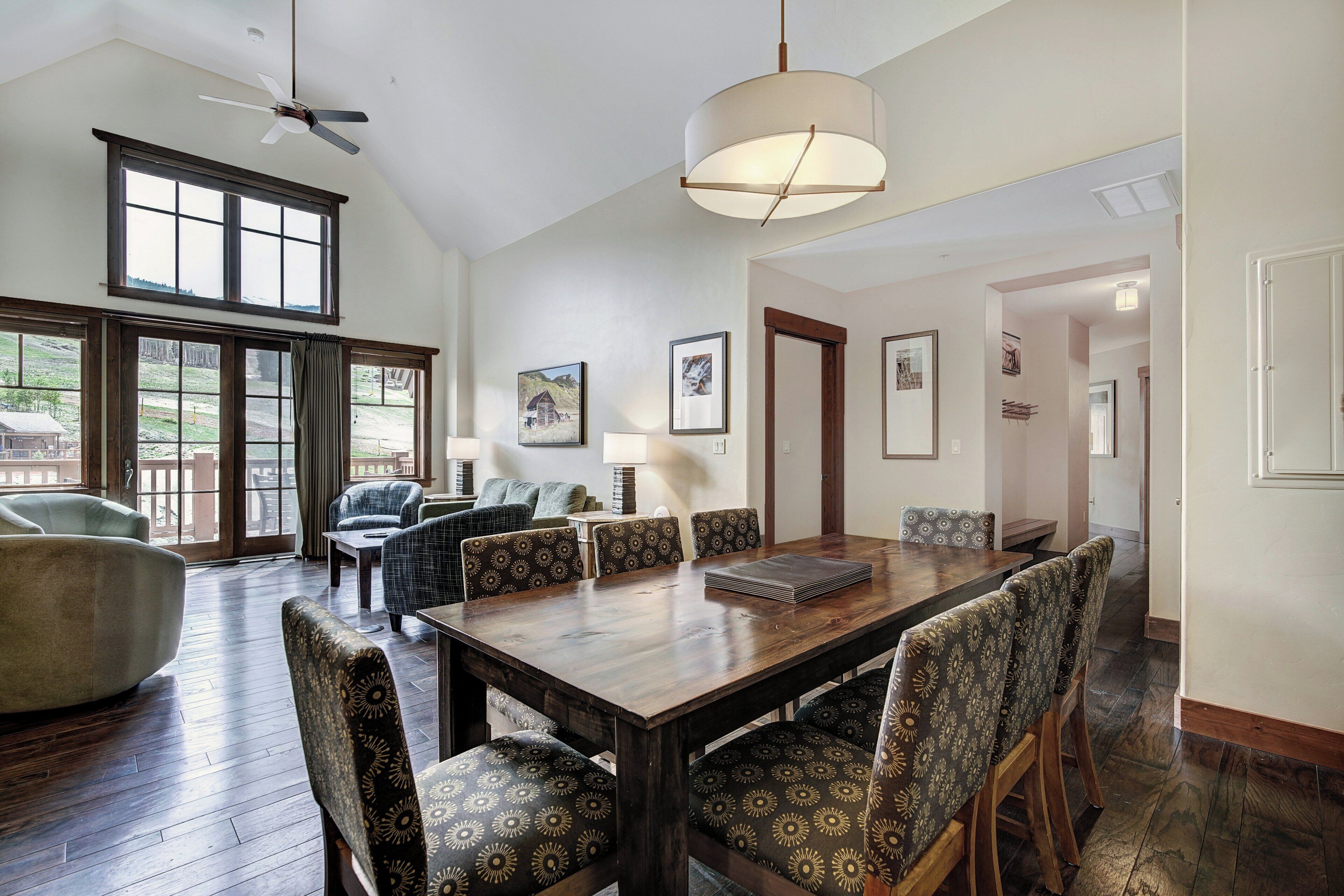 A spacious dining area with a large wooden table and patterned chairs, large windows with a view of the outdoors, and a ceiling fan.