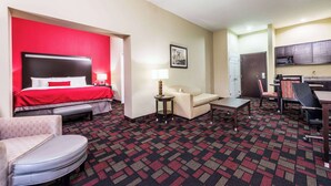 A hotel room with a red accent wall, a bed, a sofa, a desk, a chair, and a kitchenette. - Wingate by Wyndham Lake Charles Casino Area (Lake Charles)