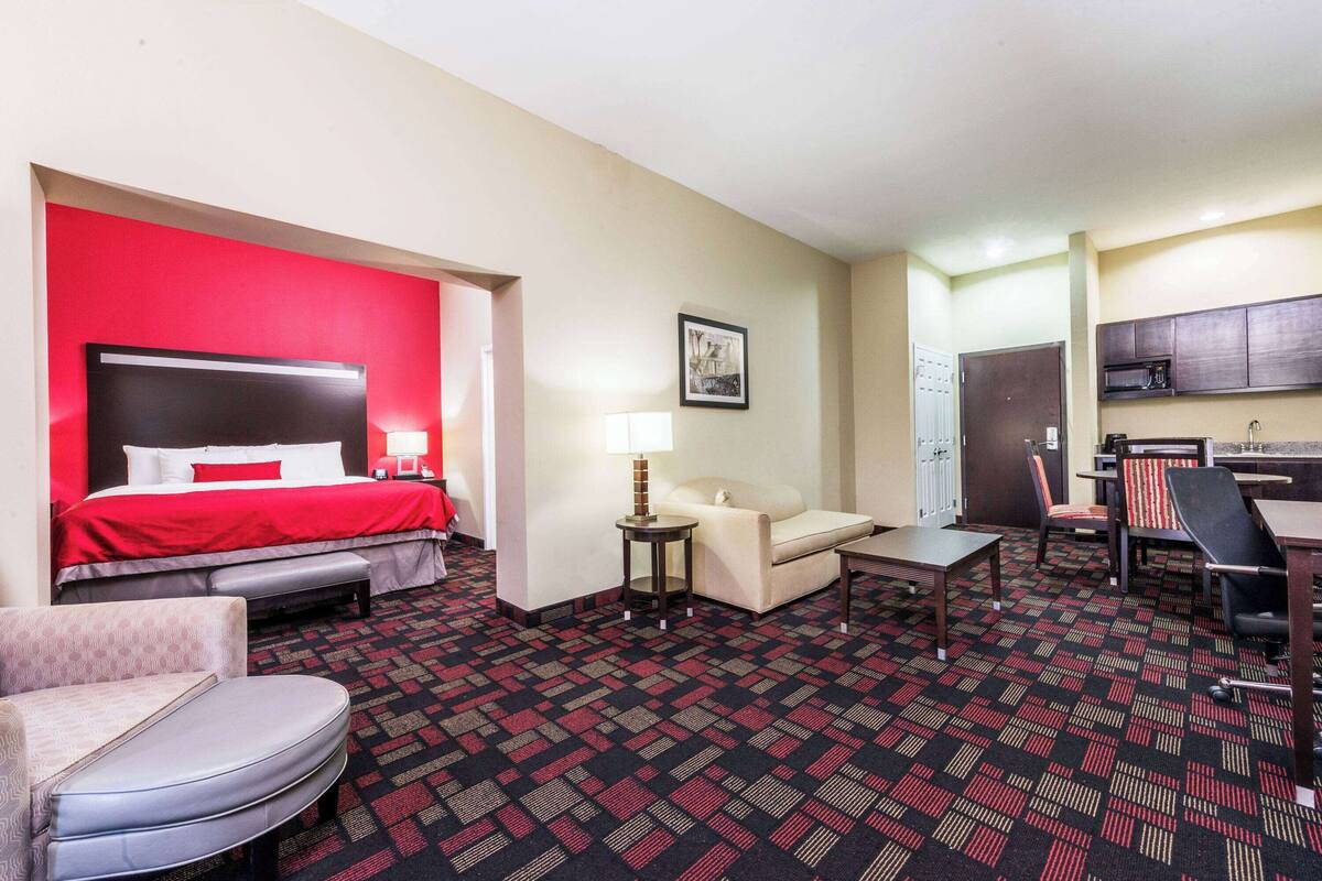 Ein Hotelzimmer mit roter Akzentwand, einem Bett, einer Couch, einem Schreibtisch, einem Sessel und einer Küchenzeile.