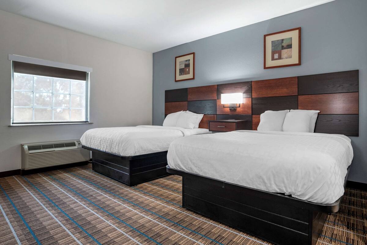 A hotel room with two beds, a window with blinds, a wall-mounted heater, and a wall-mounted lamp.