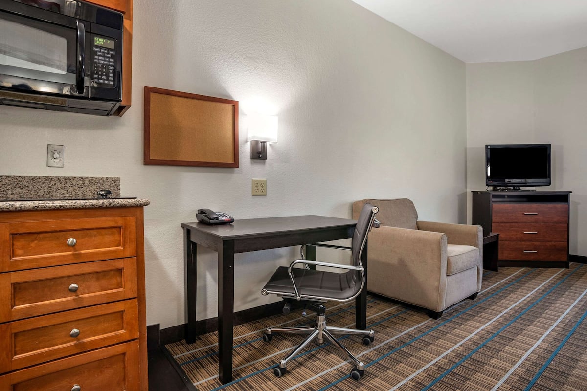 A hotel room with a kitchenette, a desk, a chair, a sofa, and a TV.