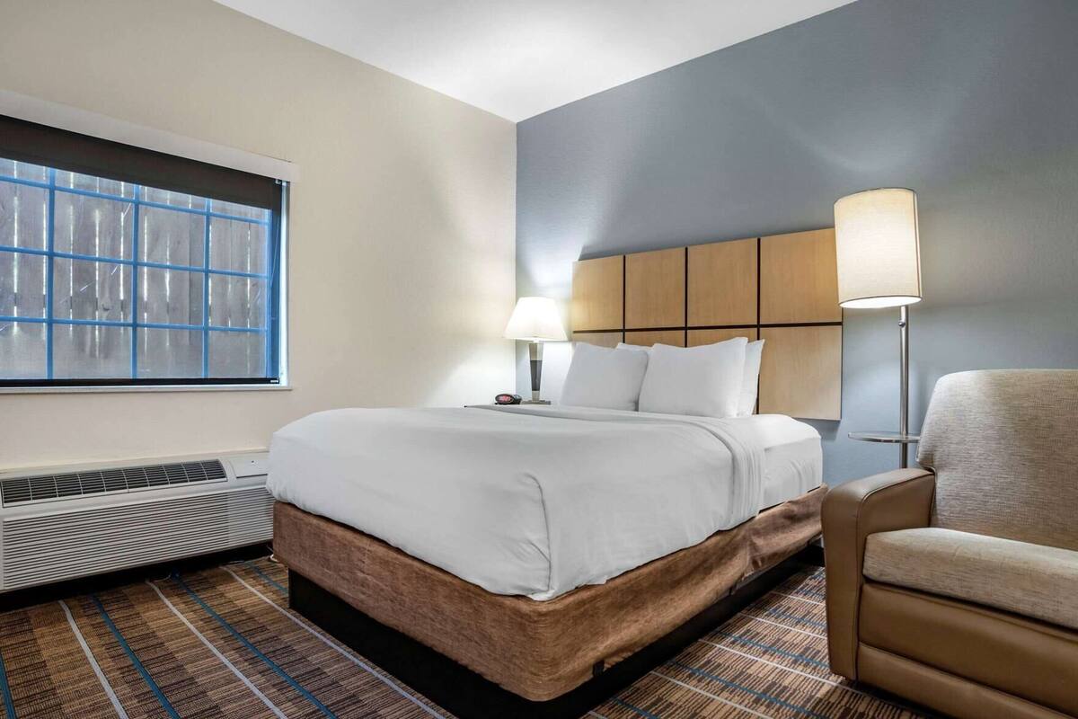 A hotel room with a bed, a chair, a window, a heater, and a lamp.