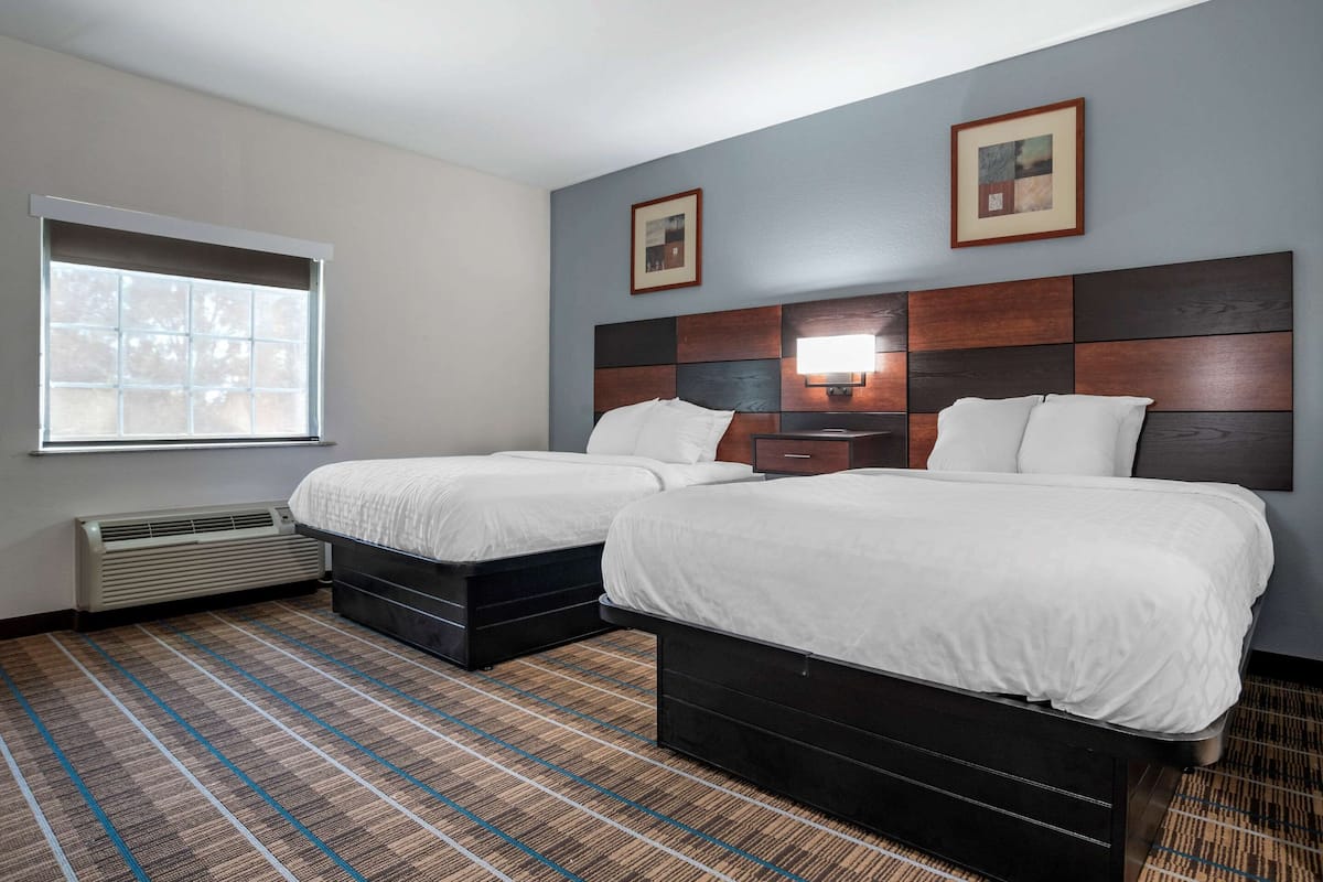 A hotel room with two beds, a large window, a wall-mounted heater, and a nightstand with a lamp.