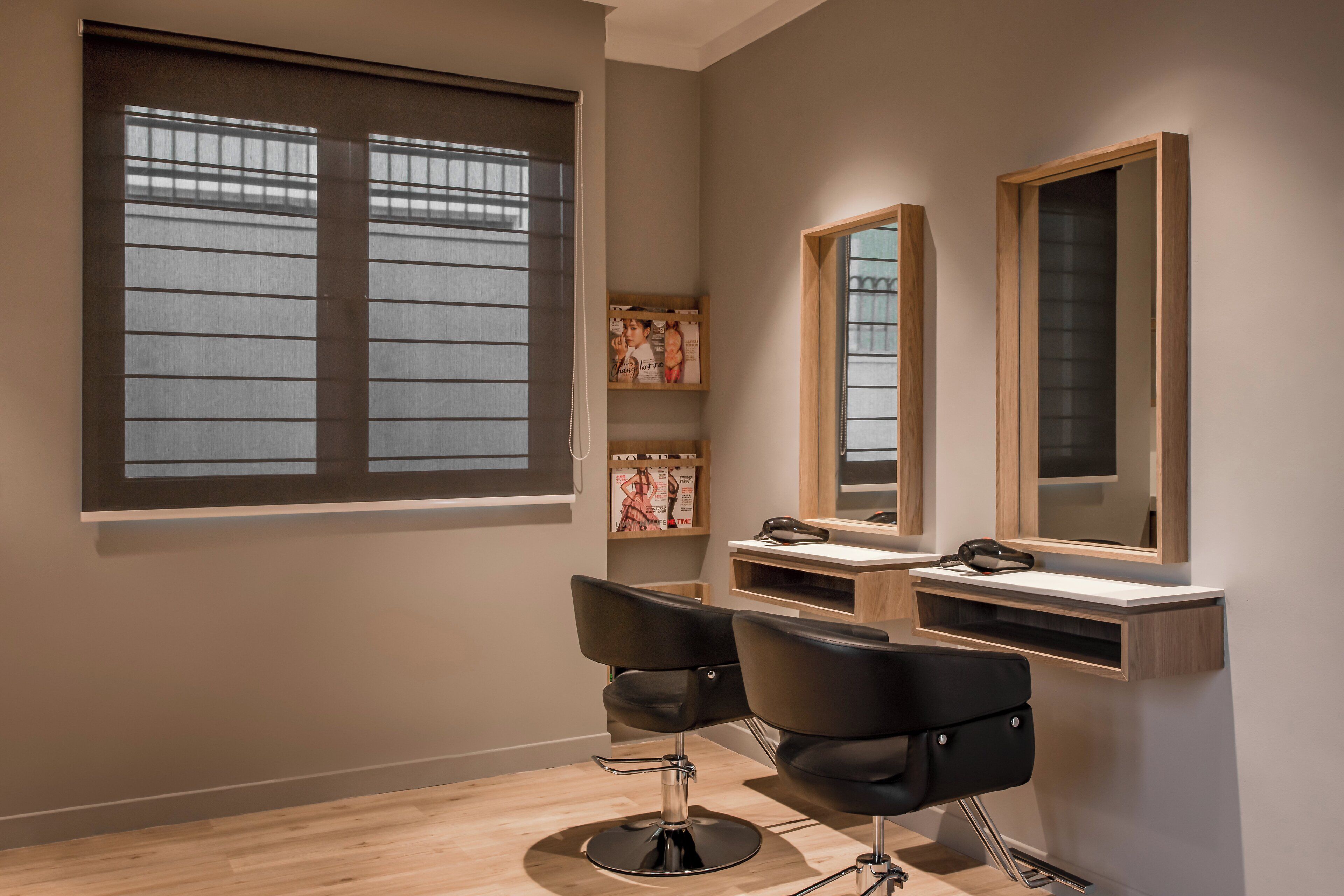 A modern salon with two black chairs, a large window with blinds, and a wooden vanity with a mirror.
