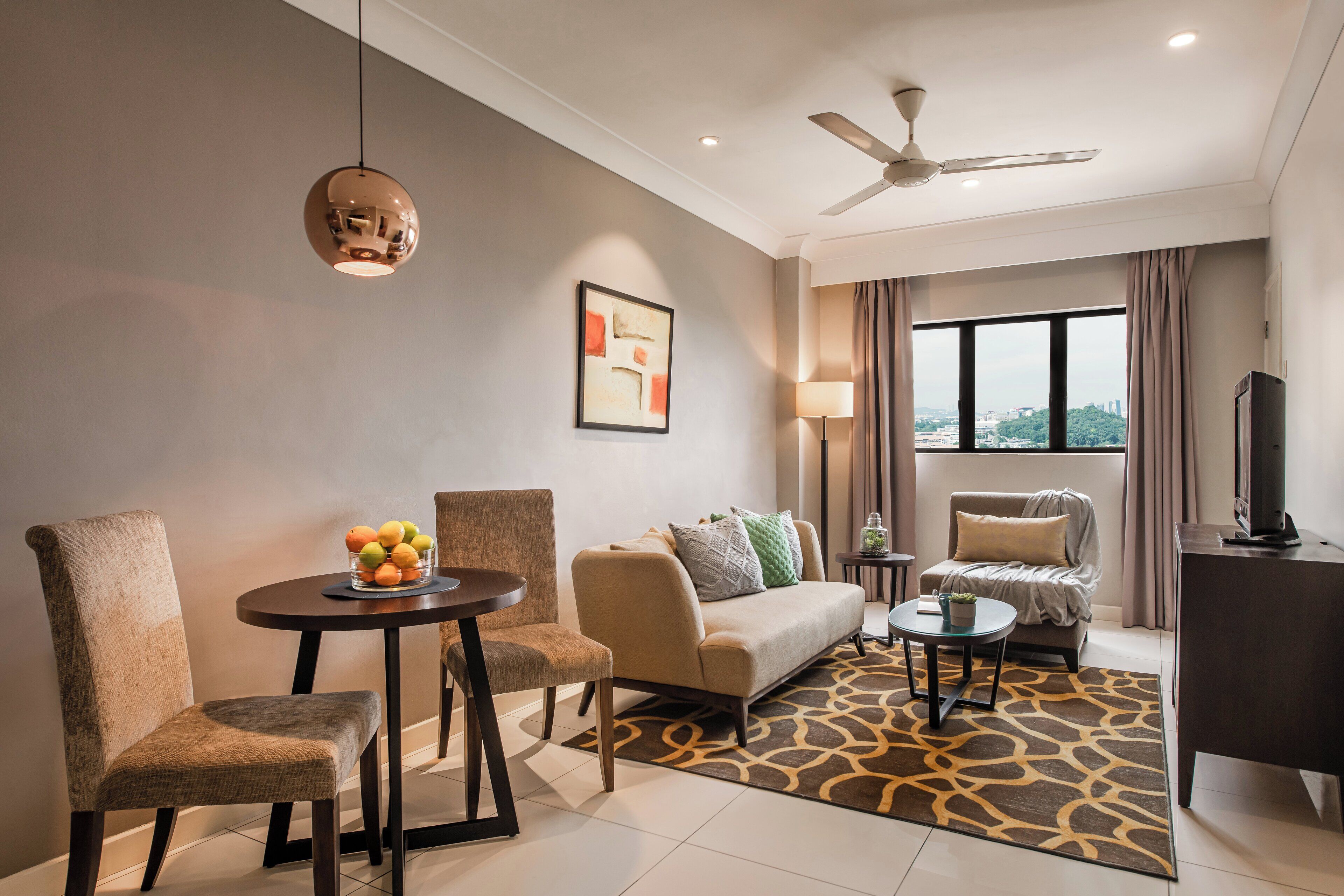 A modern hotel room with a sofa, two armchairs, a dining table with fruit, a TV, and a ceiling fan.