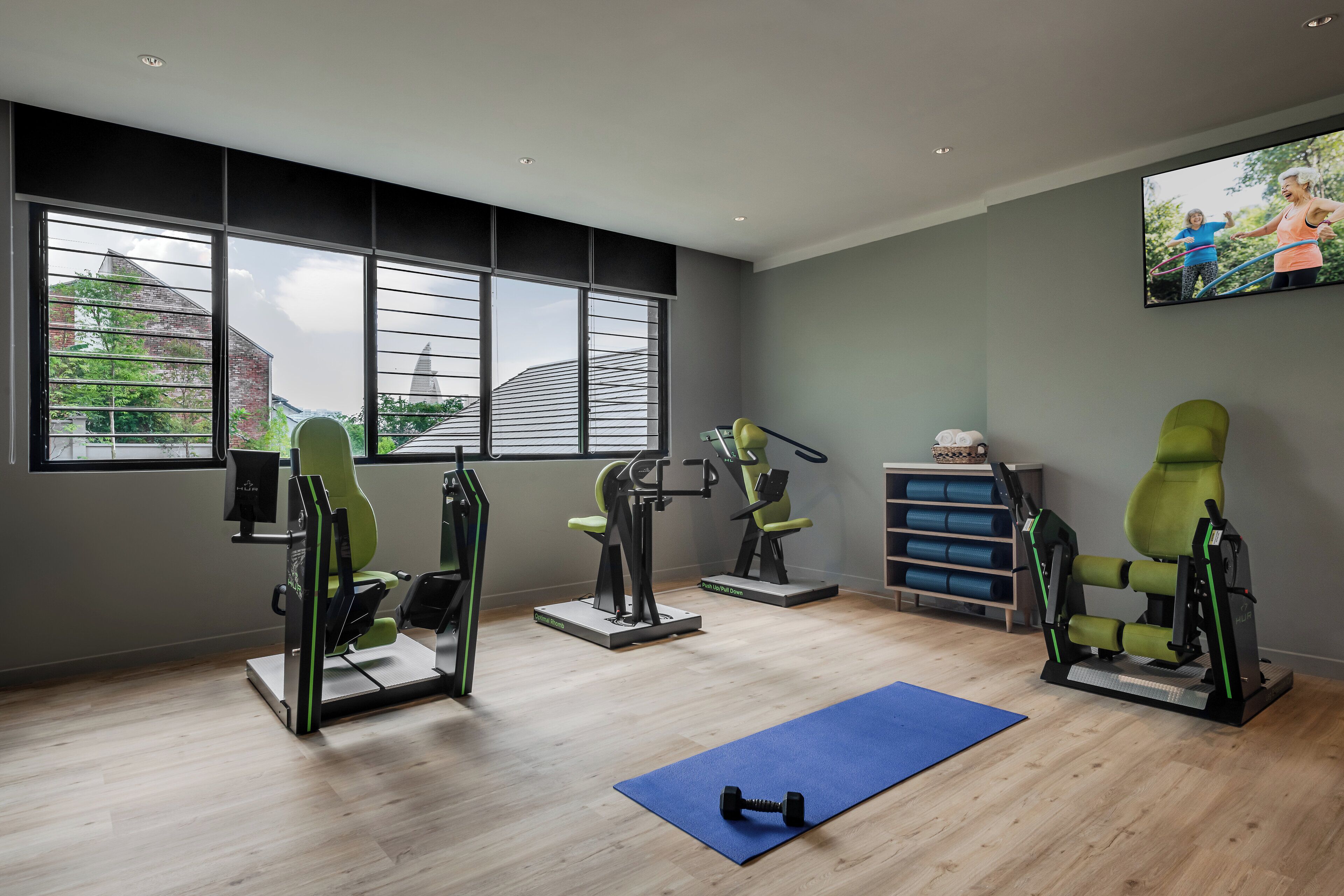 Ein Fitnessraum mit Trainingsgeräten, einer blauen Matte und einem Fernseher, der Menschen beim Training zeigt.