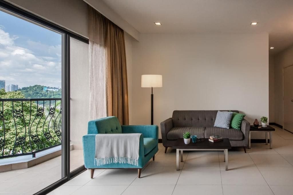 A modern living room with a sofa, armchair, and coffee table, a floor lamp, and a view of greenery through large windows.