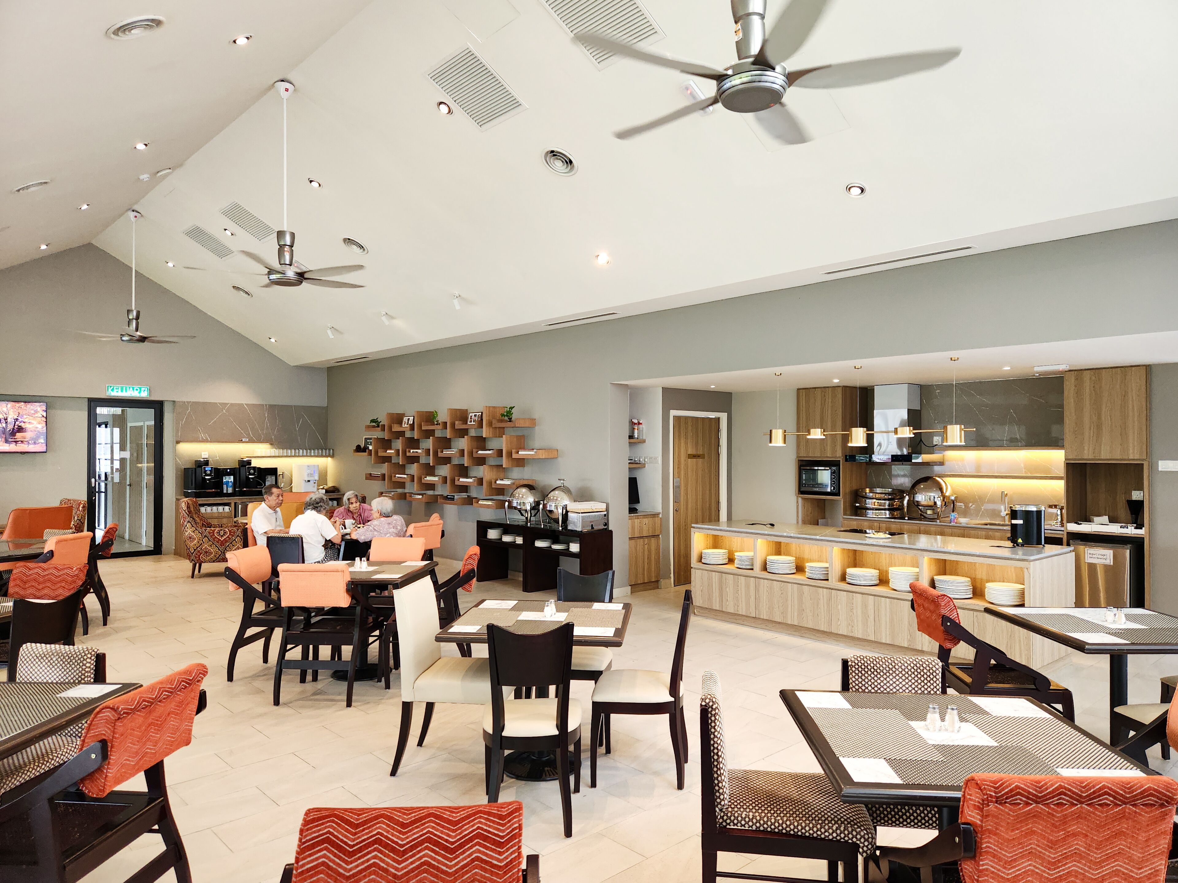 Um restaurante moderno com várias mesas e cadeiras, ventiladores de teto e uma área de cozinha visível ao fundo.