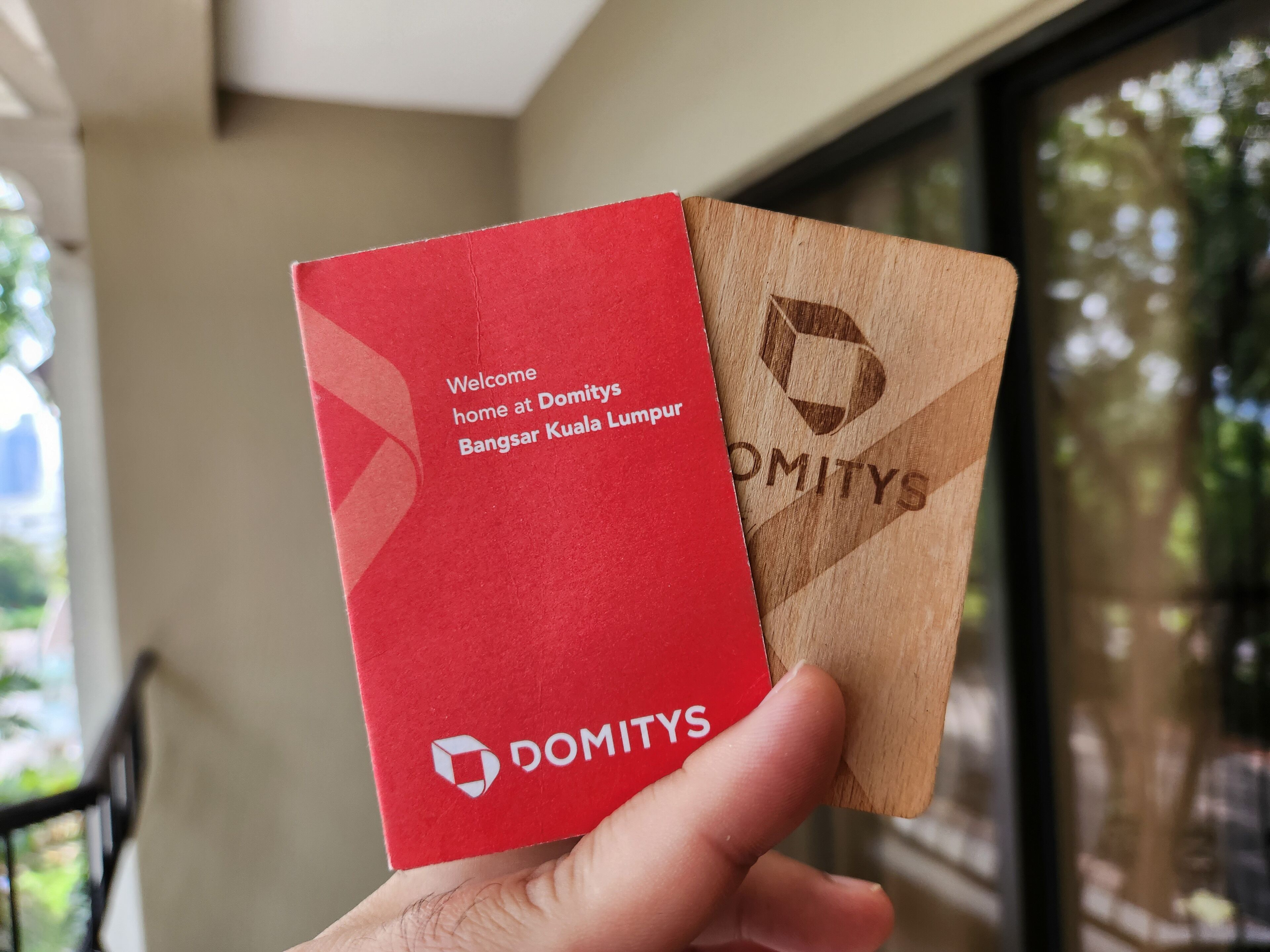 Käsi, jossa on punaruskea kortti, jossa lukee teksti: ”Tervetuloa kotiin Domitys Bangsar Kuala Lumpuriin” ja jossa on Domitysin logo.