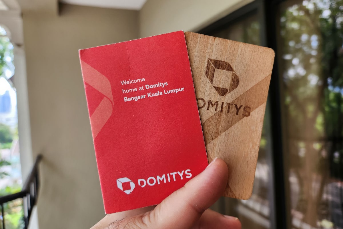 Käsi, jossa on punaruskea kortti, jossa lukee teksti: ”Tervetuloa kotiin Domitys Bangsar Kuala Lumpuriin” ja jossa on Domitysin logo.
