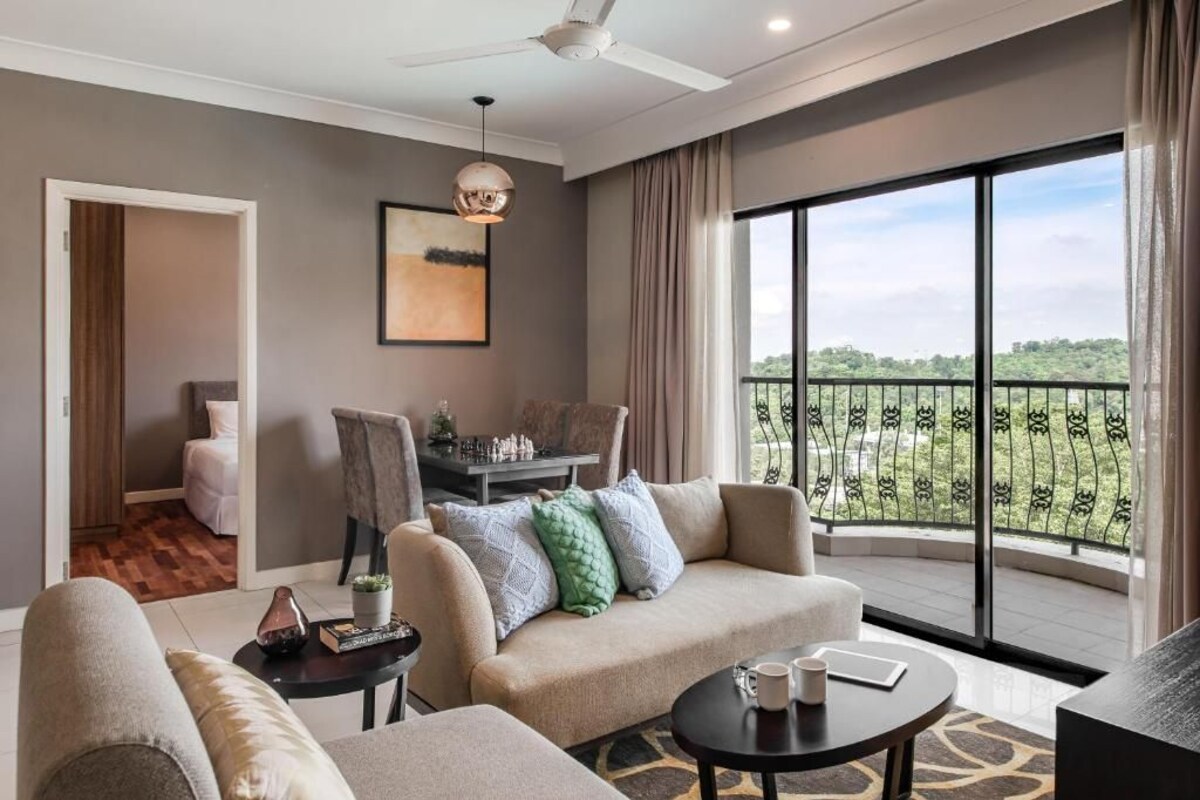 A modern living room with a sofa, armchair, and coffee table. There is a dining area with a table and chairs. The room has large windows with a view of greenery.