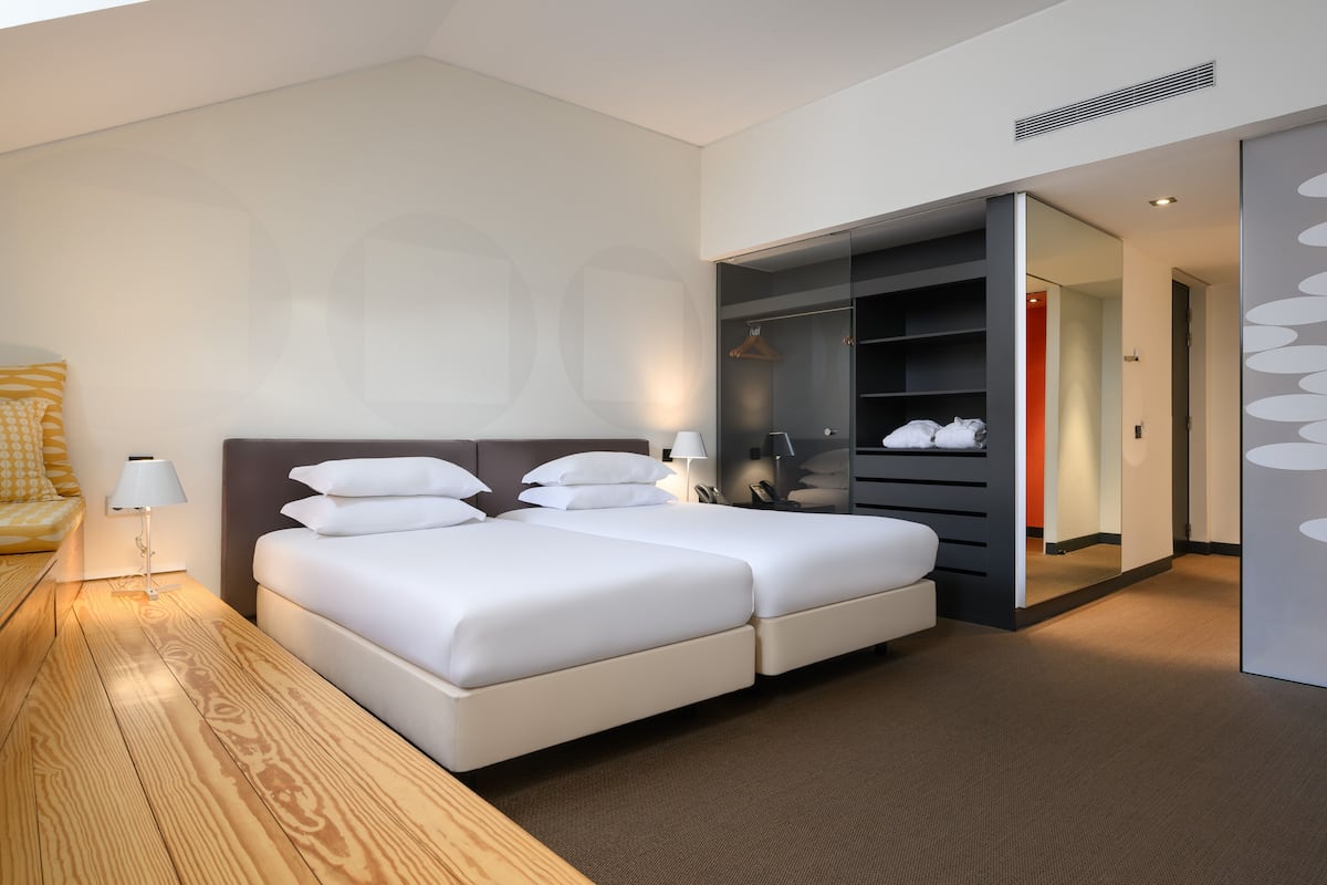 A modern hotel room with a large bed, a bedside table with a lamp, a wooden floor, and a built-in wardrobe with a mirror.