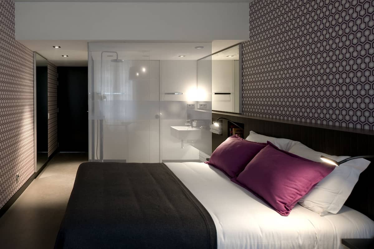 A modern hotel room with a large bed, a bedside table, a mirror, a shower area, and a patterned wall.