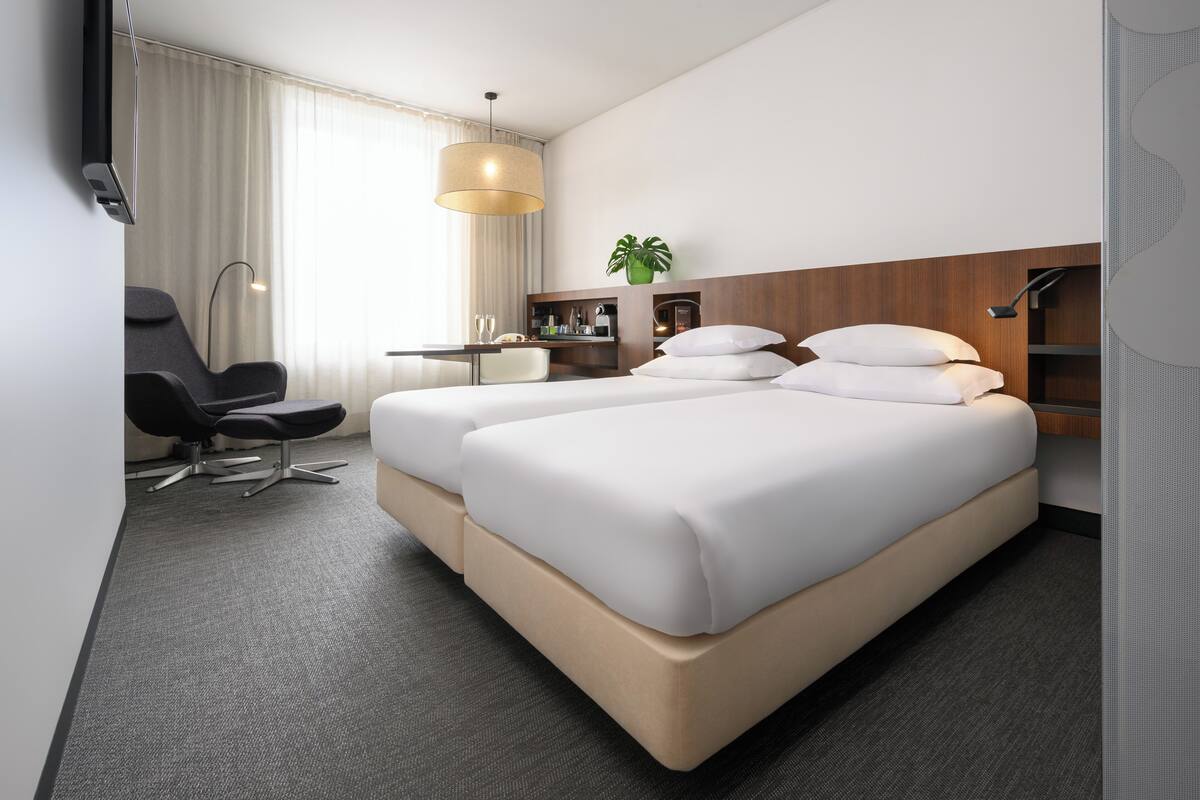 A hotel room with a large bed, a desk, a chair, a plant, and a hanging light fixture.