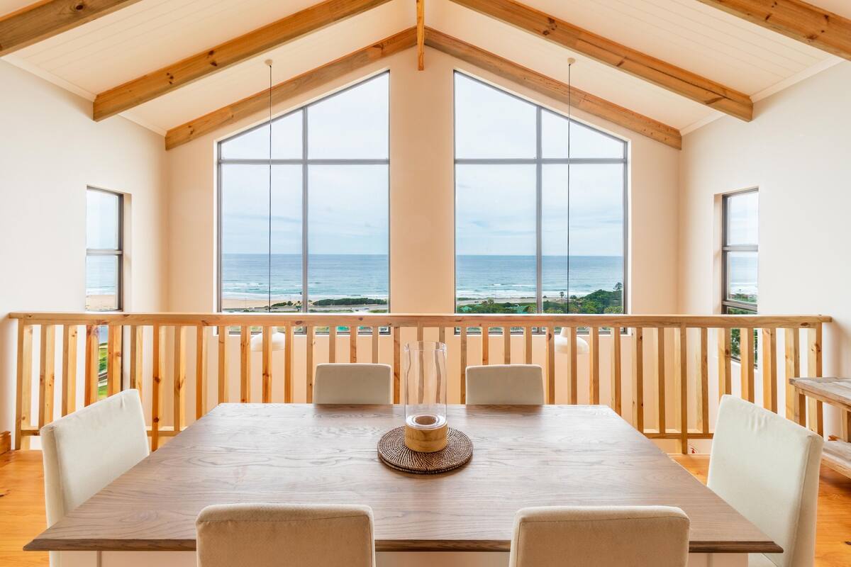 A dining area with a wooden table and chairs, large windows with a view of the ocean, and a wooden balcony railing.