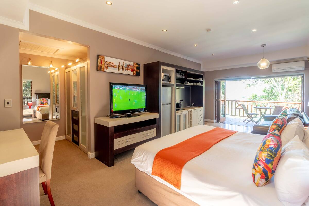 A hotel room with a bed, a TV, a desk, and a balcony with outdoor furniture.