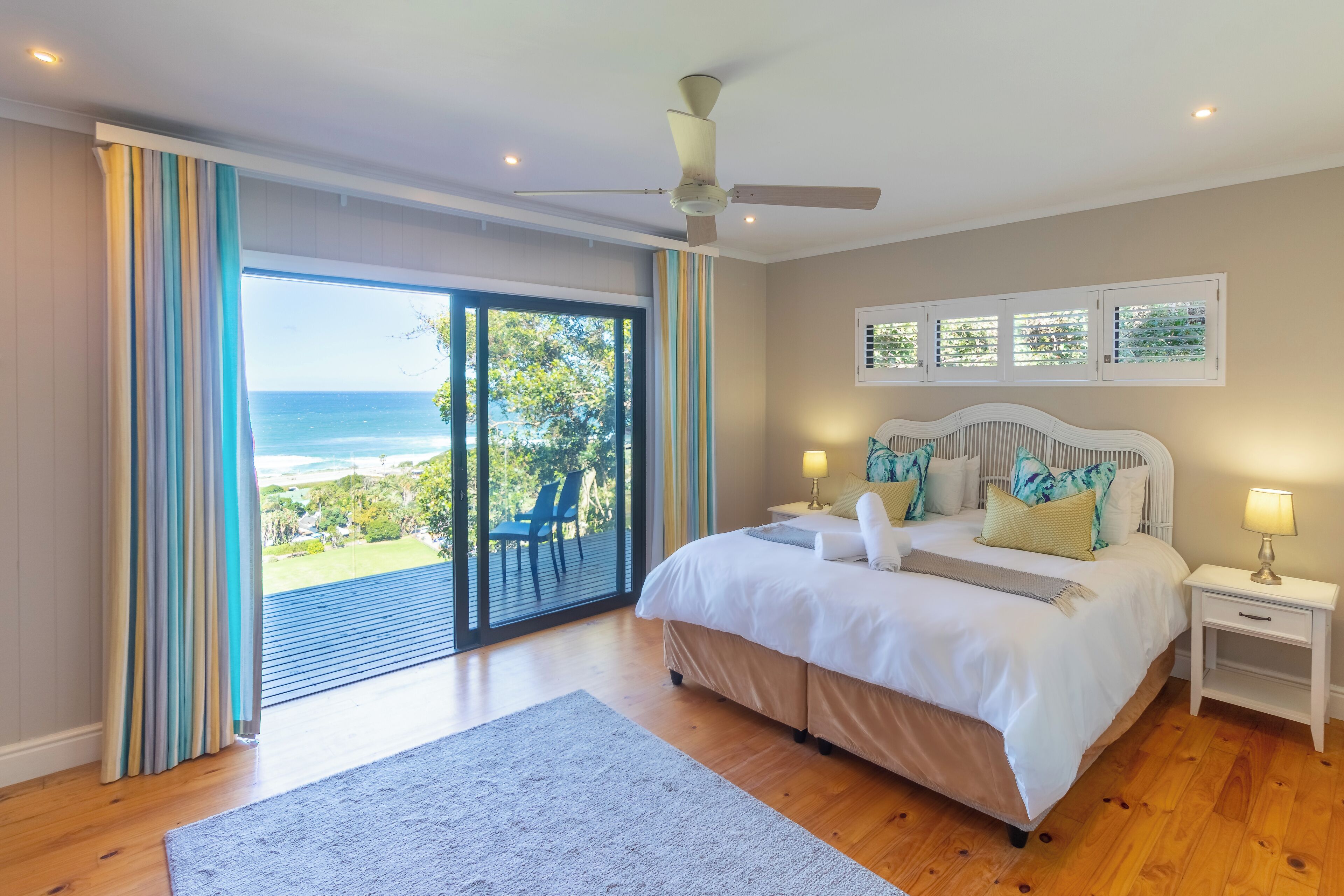 A bedroom with a large bed, a ceiling fan, a nightstand with a lamp, and a view of the ocean through a sliding door.
