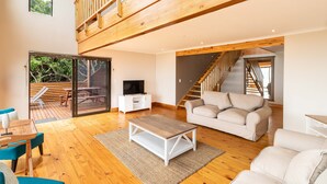 A spacious living area with a wooden floor, a white sofa, a coffee table, and a TV on a white cabinet. There is a staircase leading to an upper level. - Boardwalk Lodge - self-catering (Wilderness)