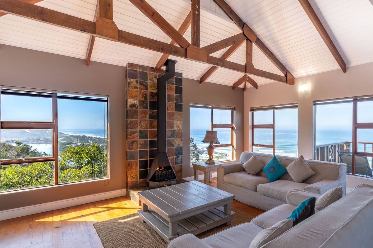 A spacious living room with a stone fireplace, large windows offering ocean views, and a high ceiling with exposed beams.